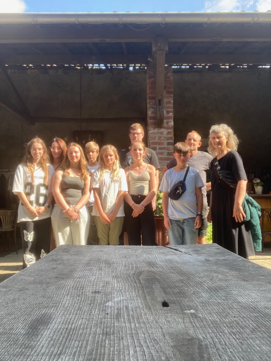 Gruppe von neun Personen steht vor einer Backsteinwand unter einem überdachten Bereich an einem sonnigen Tag