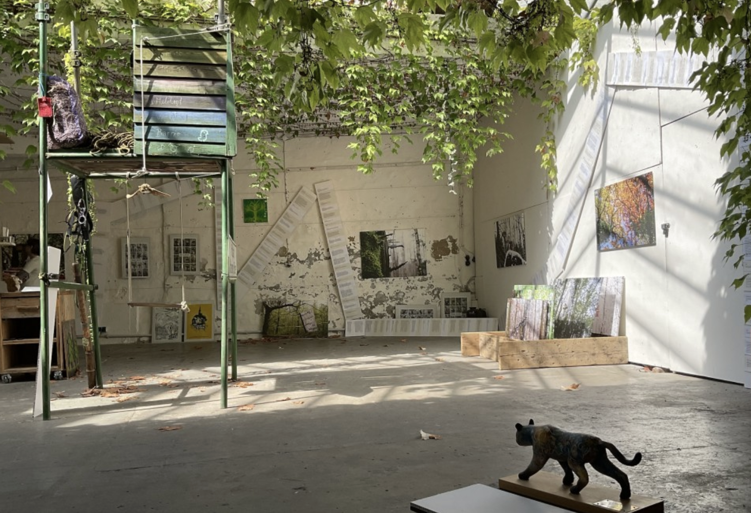 Blick in die Ausstellungsfläche mit Hochsitzobjekt und Fotos an einer Timeline ausgerichtet, Figur einer schwarzen Katze im Vordergrund.