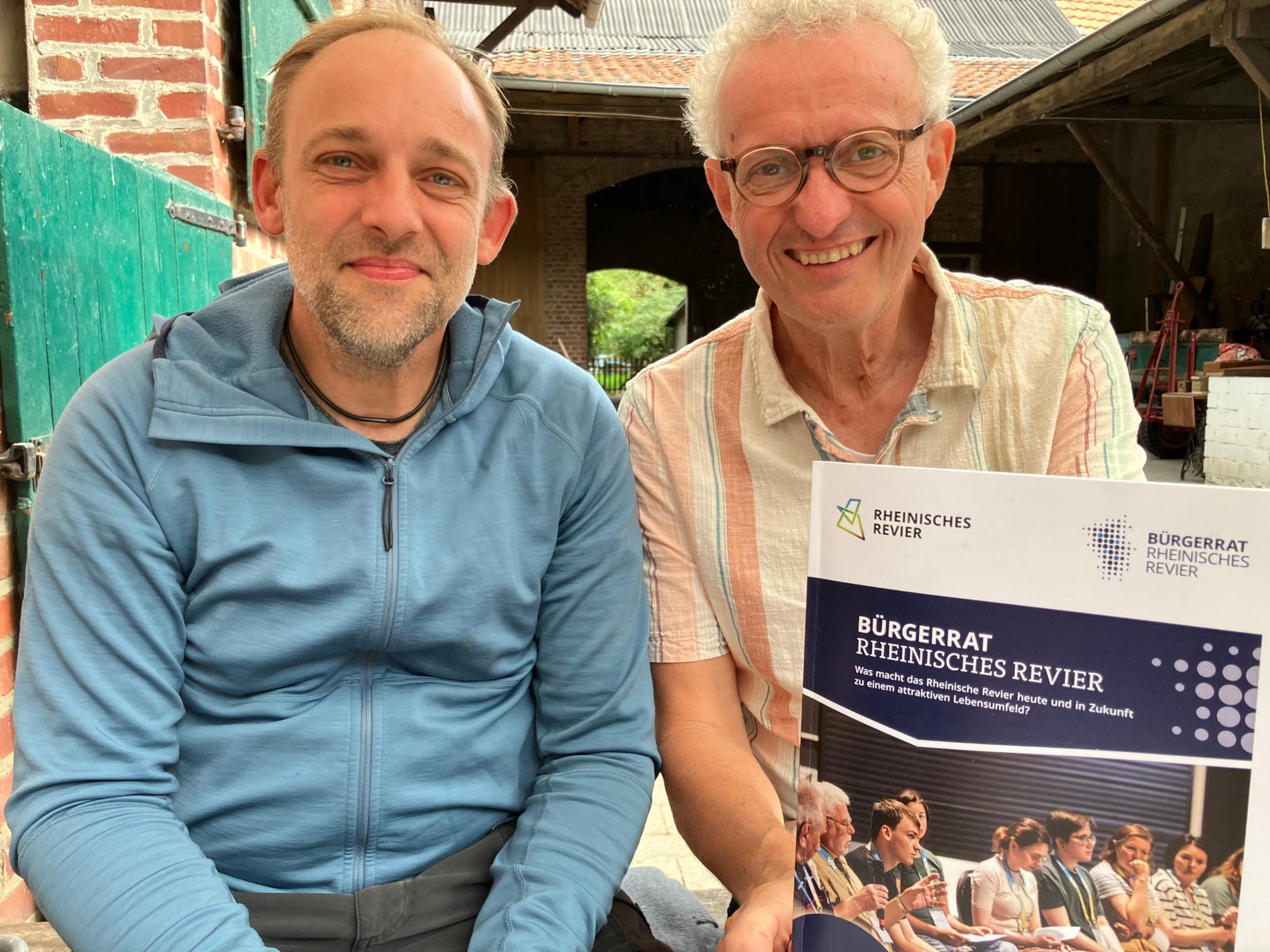 Zwei Personen sitzen nebeneinander, einer hält ein Plakat mit der Überschrift 'BÜRGERRAT RHEINISCHES REVIER'