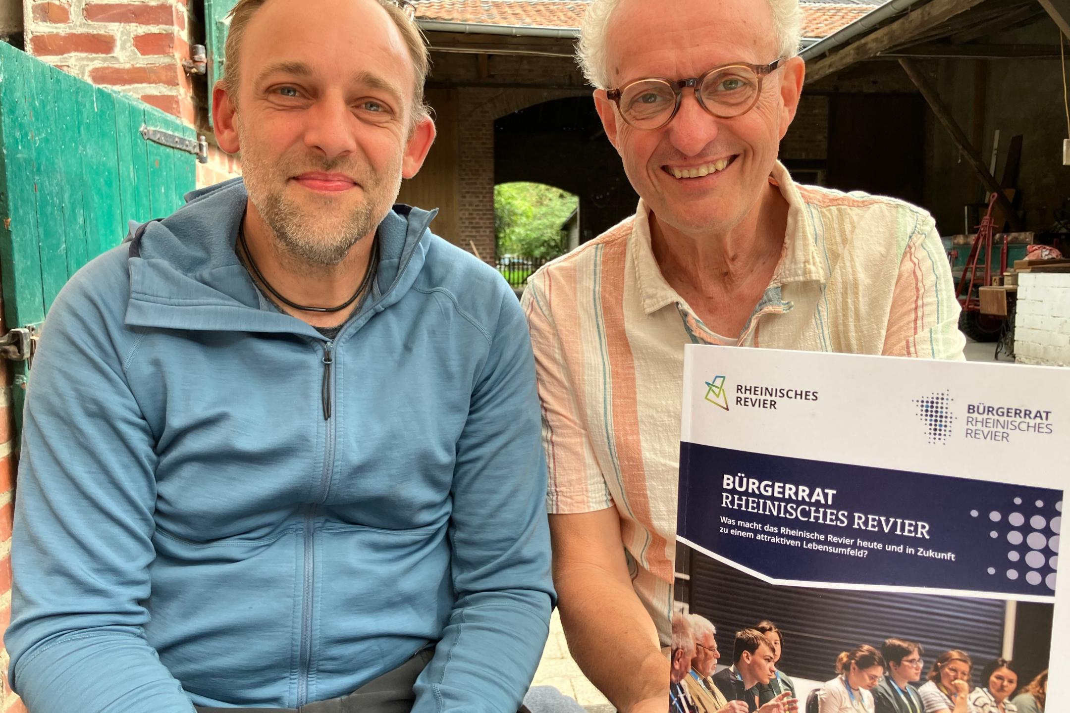 Zwei Personen sitzen nebeneinander, einer hält ein Plakat mit der Überschrift 'BÜRGERRAT RHEINISCHES REVIER'