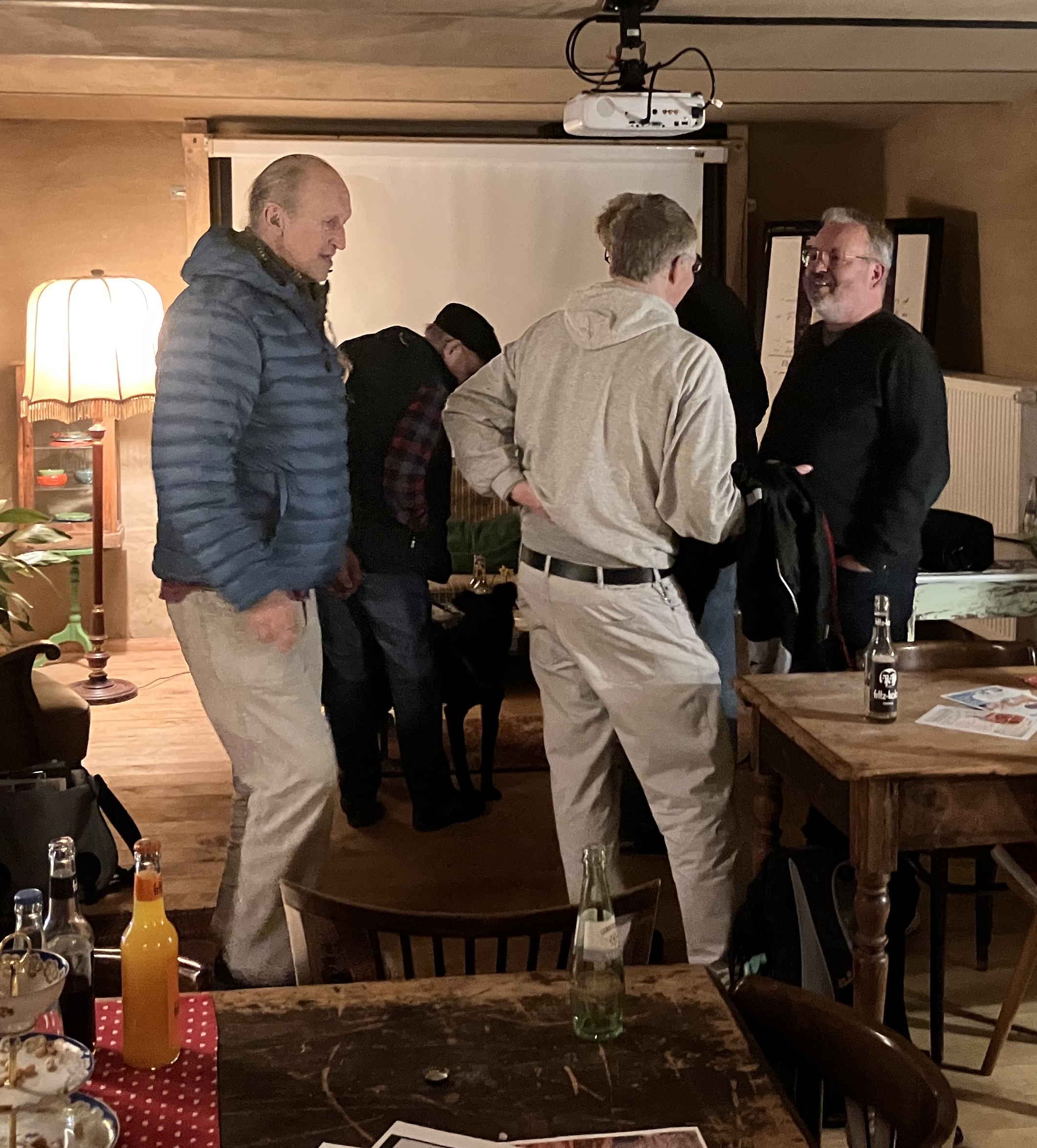 Gruppe von Männern in legerer Kleidung steht in einem Raum mit Holzmöbeln und einer Stehlampe und unterhält sich