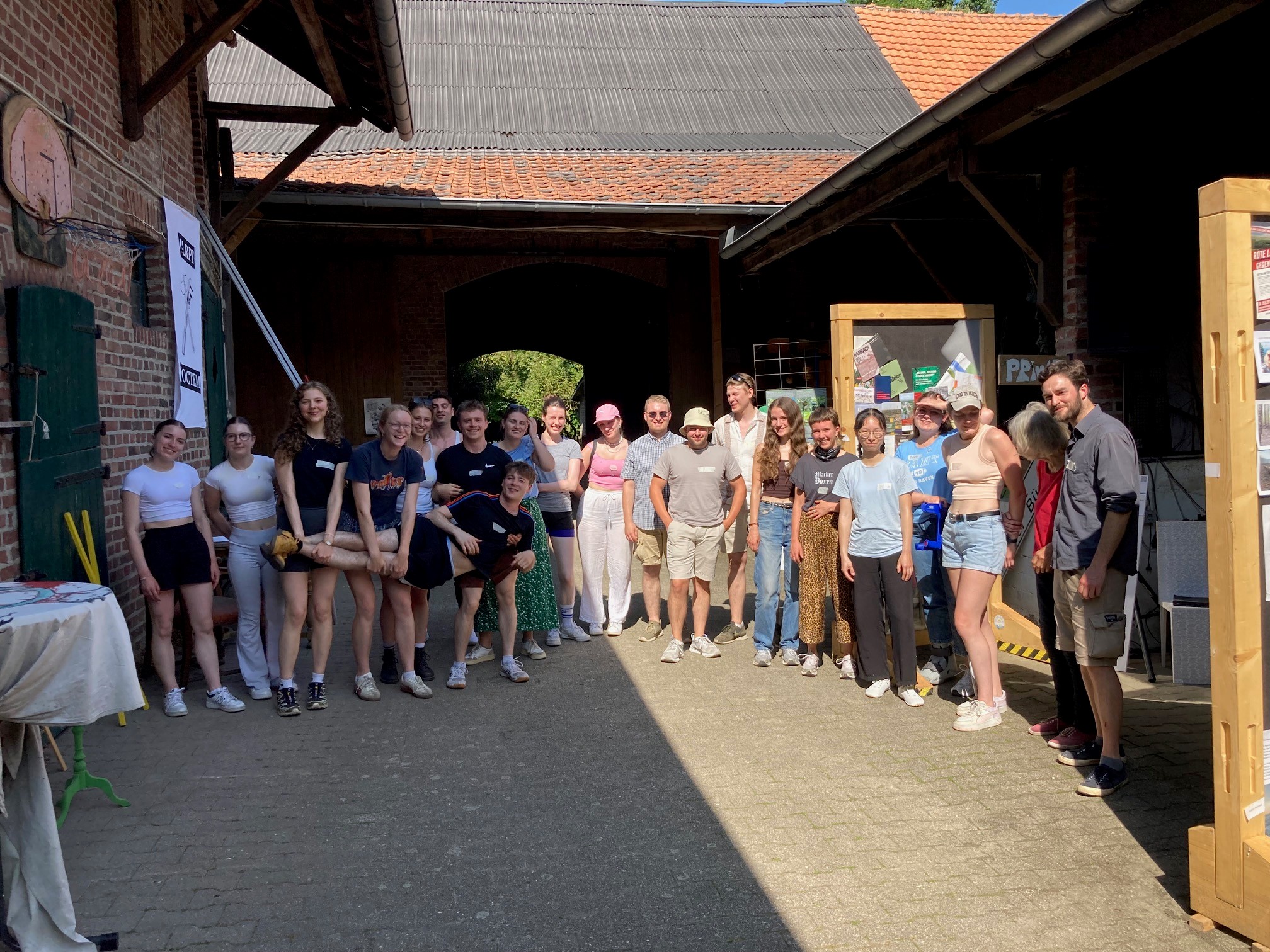 Große Gruppe junger Menschen steht in einem Hof mit Backsteinmauern und roten Dächern, einige tragen Sommerkleidung und lächeln in die Kamera