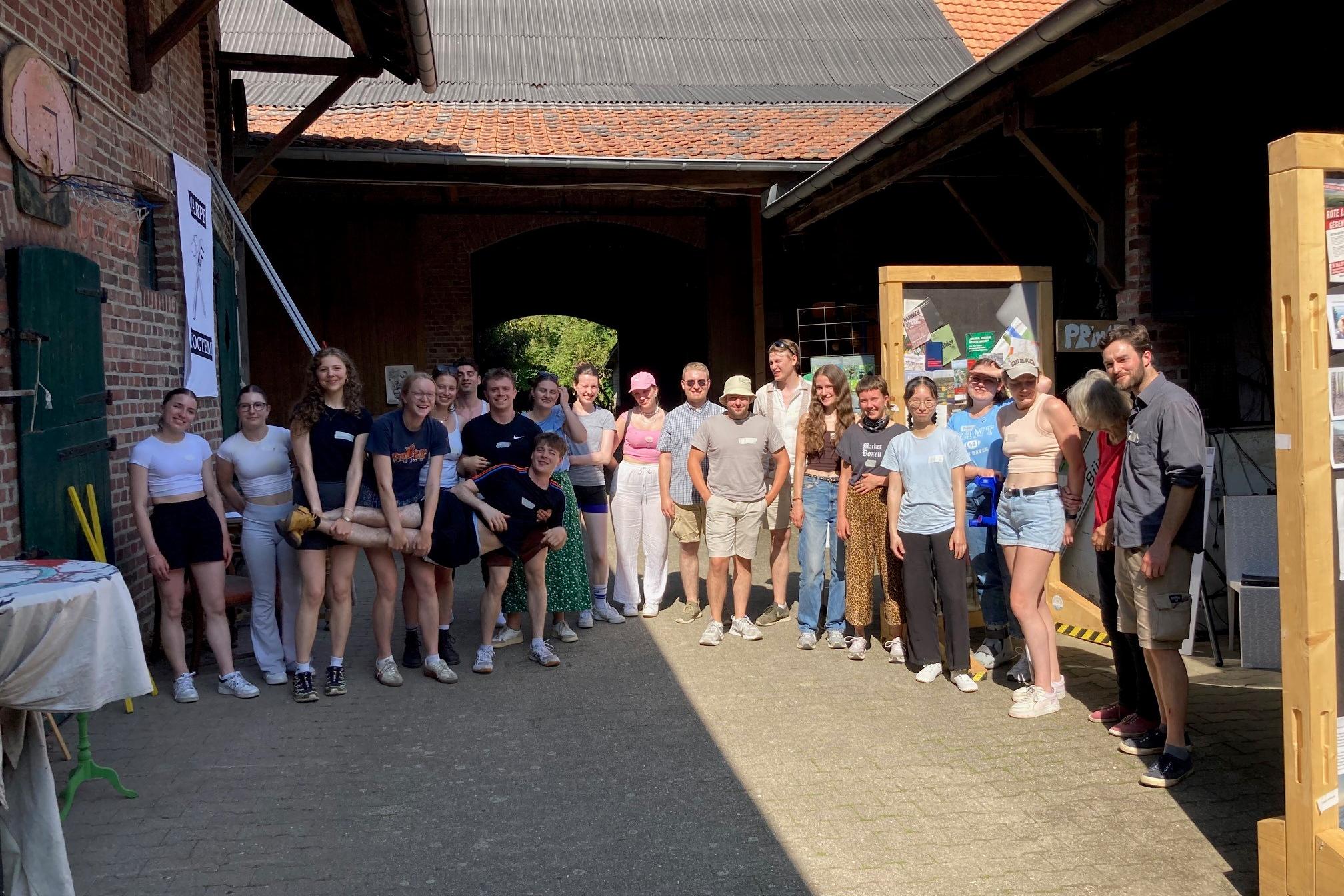 Große Gruppe junger Menschen steht in einem Hof mit Backsteinmauern und roten Dächern, einige tragen Sommerkleidung und lächeln in die Kamera