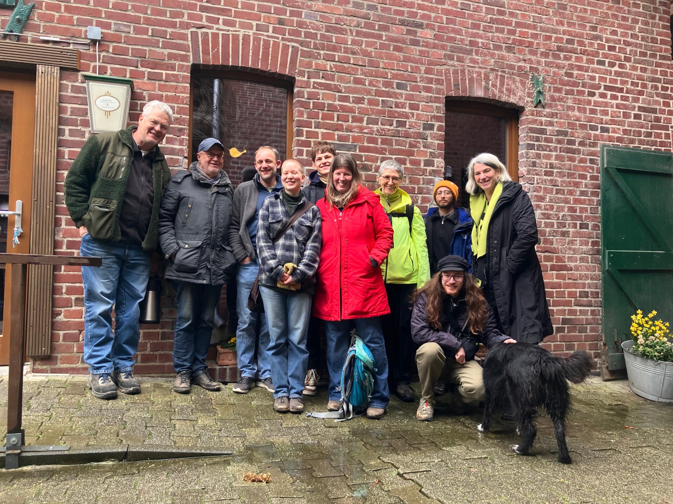 Gruppe von Menschen in Outdoor-Kleidung vor roter Backsteinwand mit zwei Fenstern und grünem Türflügel, eine Person hockt neben schwarzem Hund