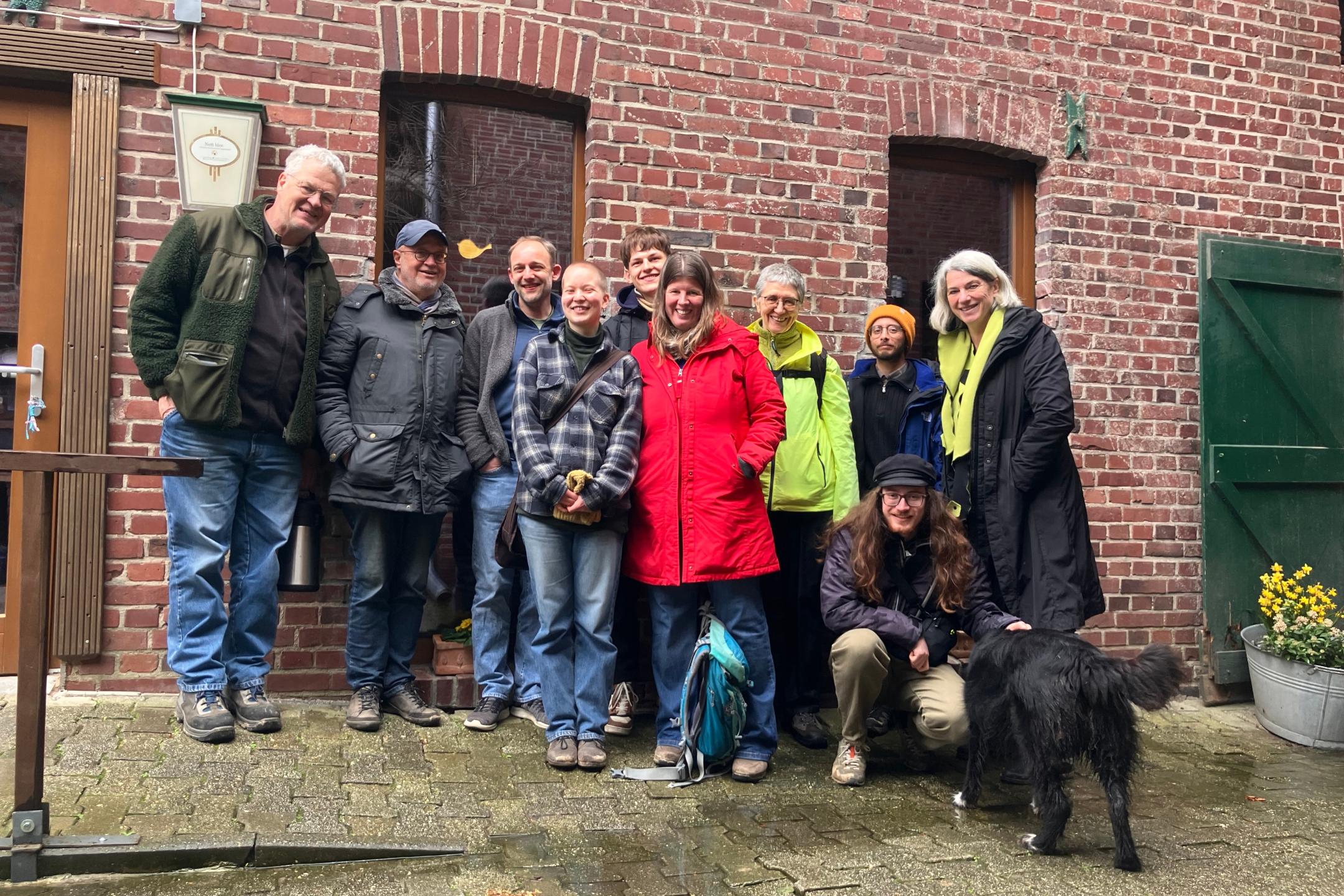 Gruppe von Menschen in Outdoor-Kleidung vor roter Backsteinwand mit zwei Fenstern und grünem Türflügel, eine Person hockt neben schwarzem Hund