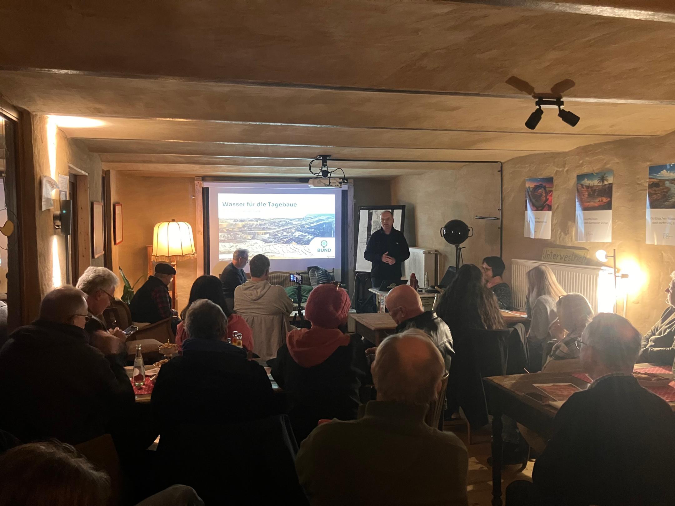 Mehrere Personen sitzen in einem Raum und hören einem Vortrag vor einer Leinwand mit der Überschrift 'Wasser für die Tagebaue' zu