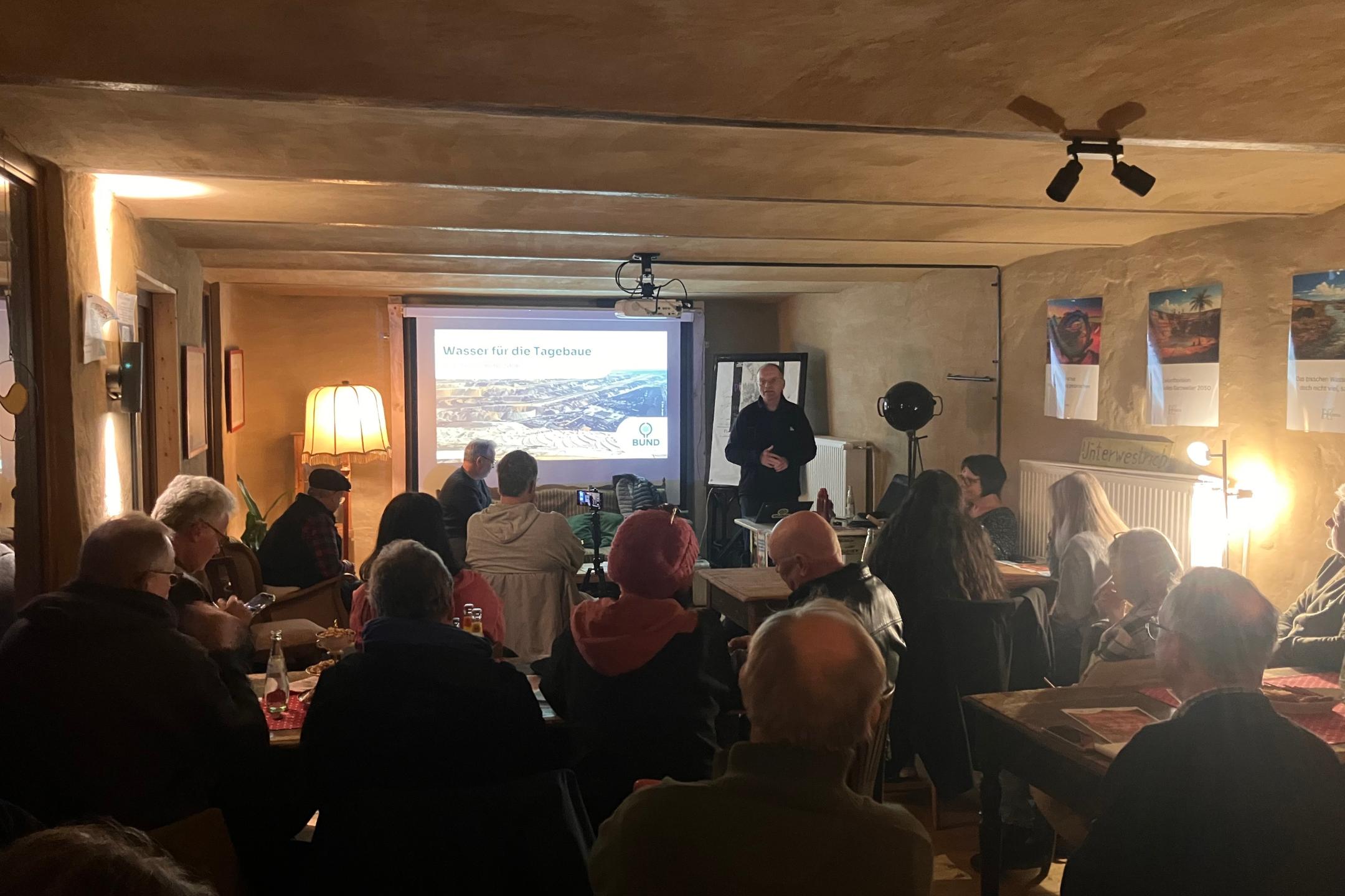 Mehrere Personen sitzen in einem Raum und hören einem Vortrag vor einer Leinwand mit der Überschrift 'Wasser für die Tagebaue' zu